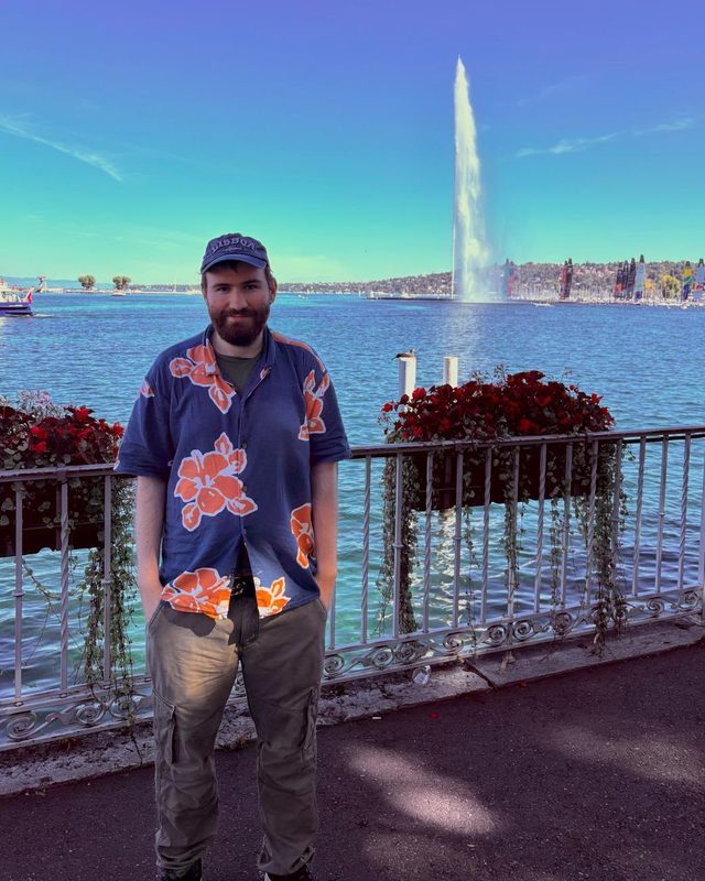 Adam Rowan standing by Lake Geneva with the Jet d'Eau fountain behind him